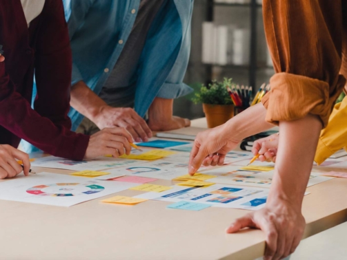 Product Marketing Process team collaborating around a table with charts, sticky notes, and planning documents during a real-world strategy session.
